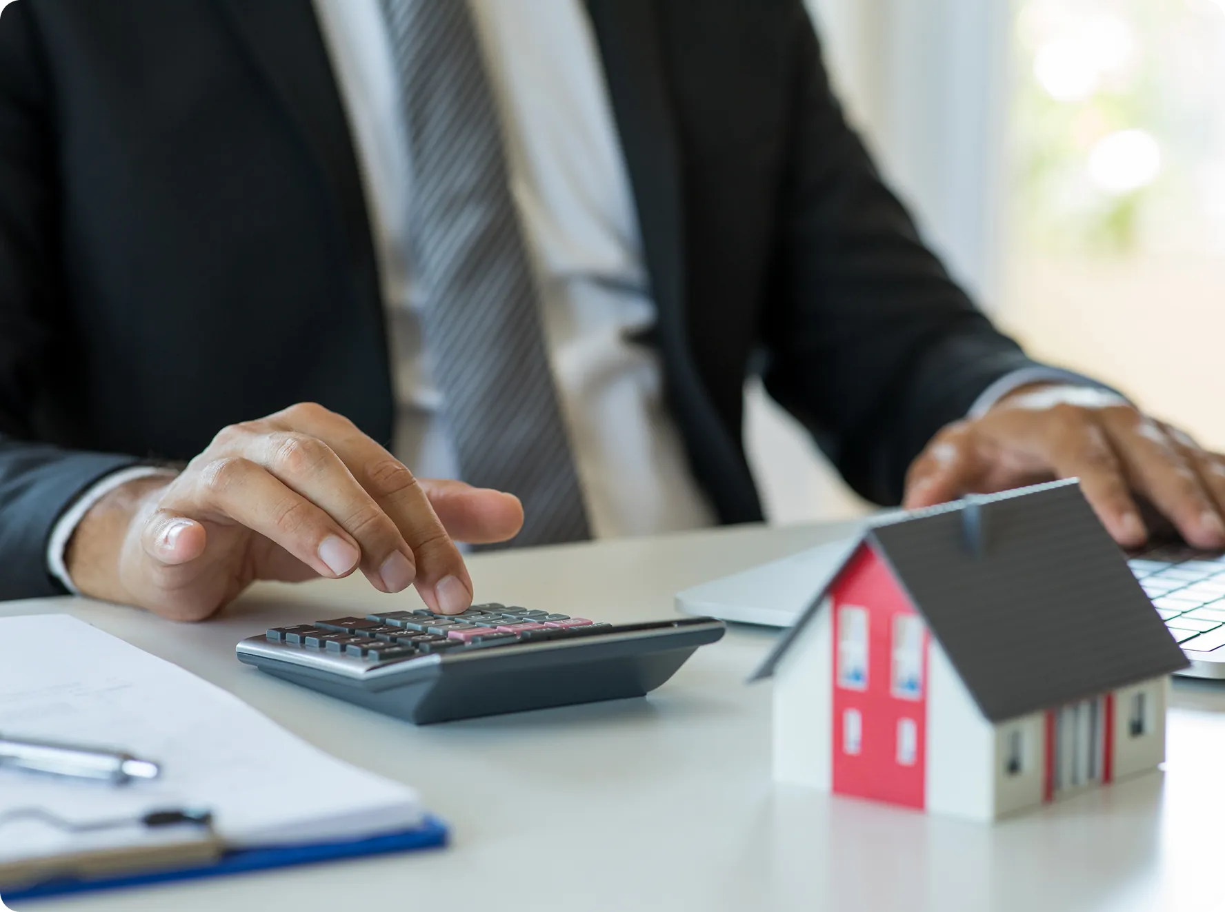 A person in a suit uses a calculator at a desk with a laptop, paper, and a small model house.