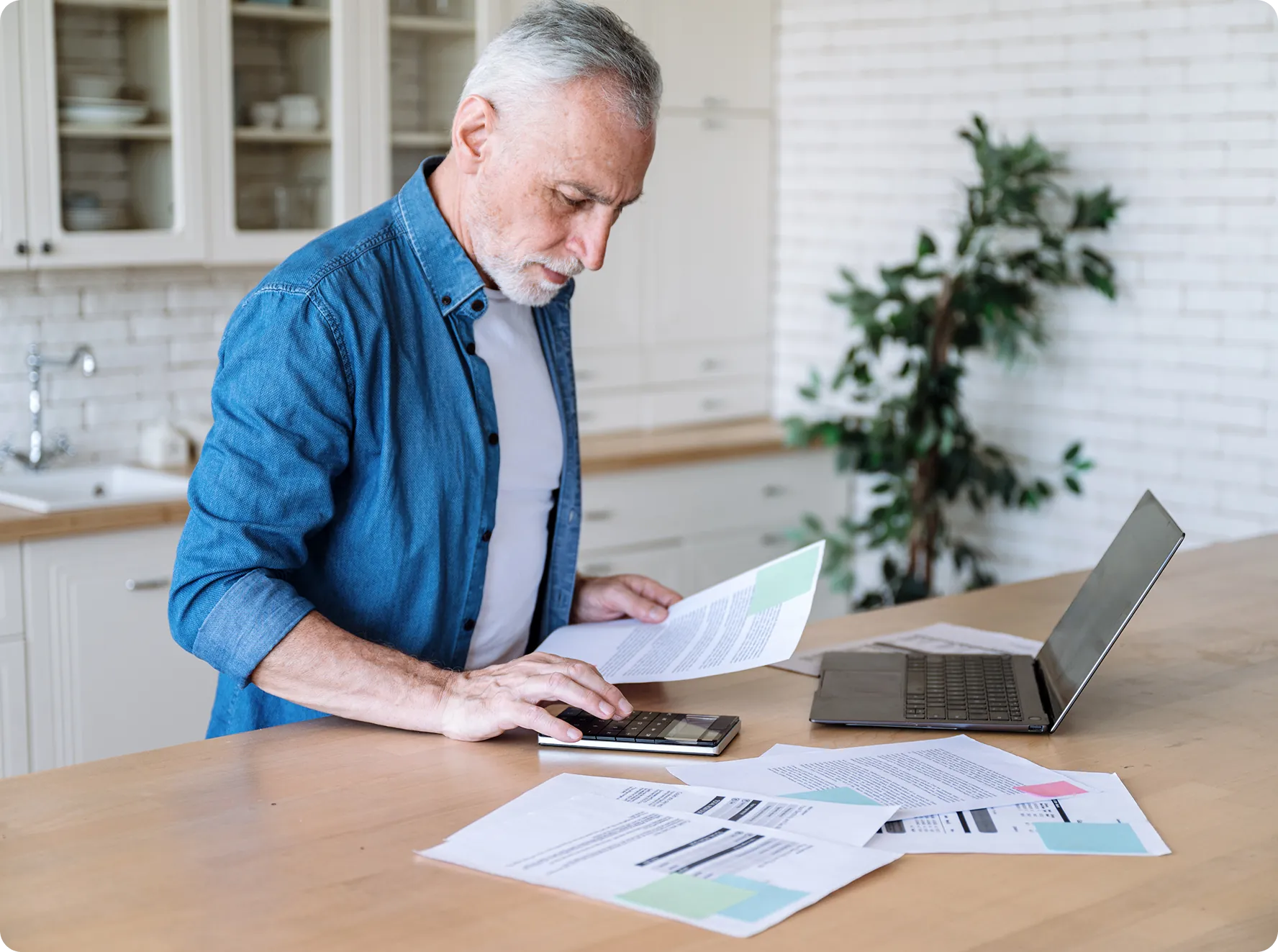 An older man stands at a kitchen counter using a calculator, with documents spread out and an open laptop beside him.