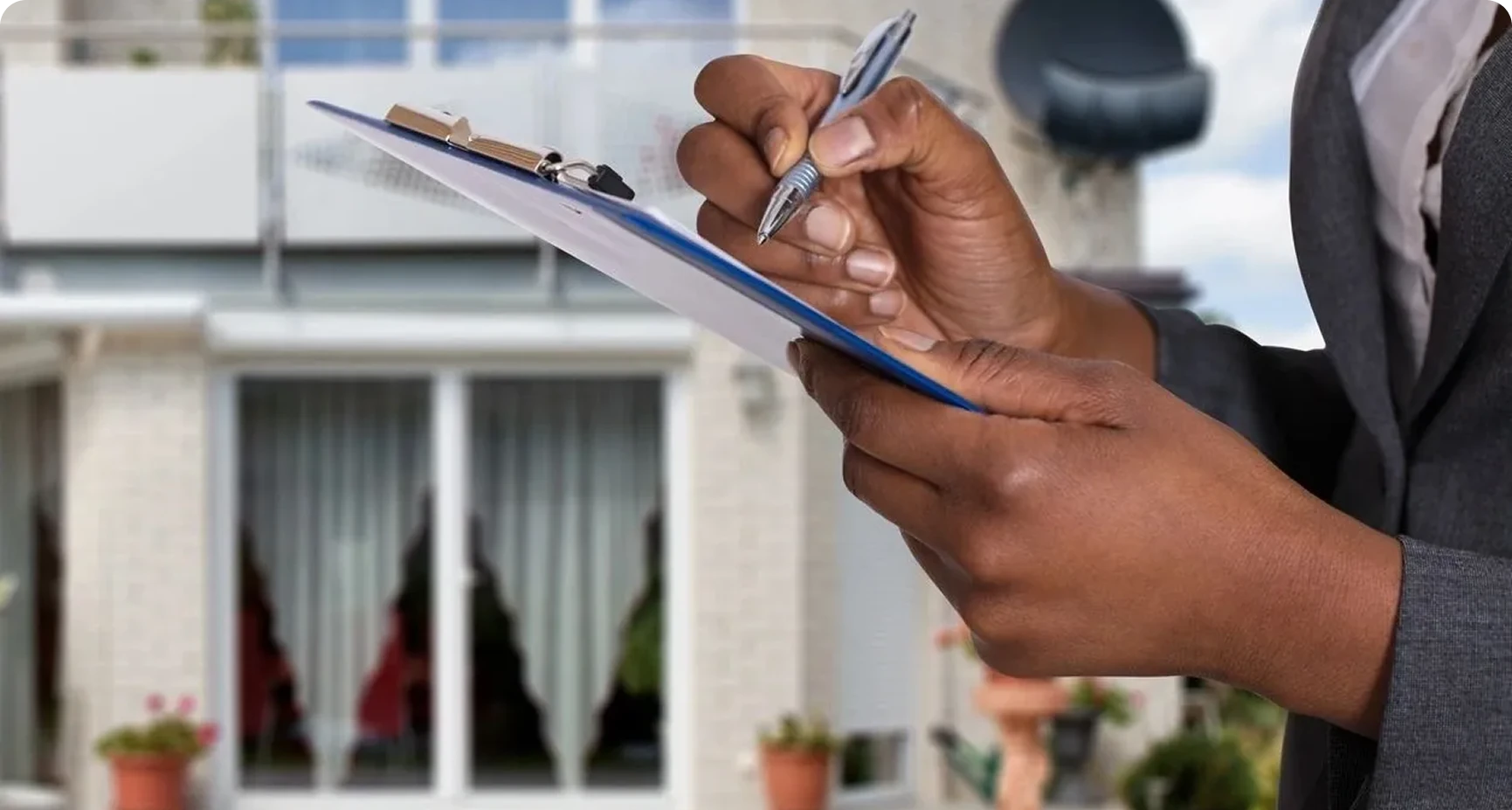 A person in business attire writes on a clipboard outside a residential building with glass doors and a balcony in the background.