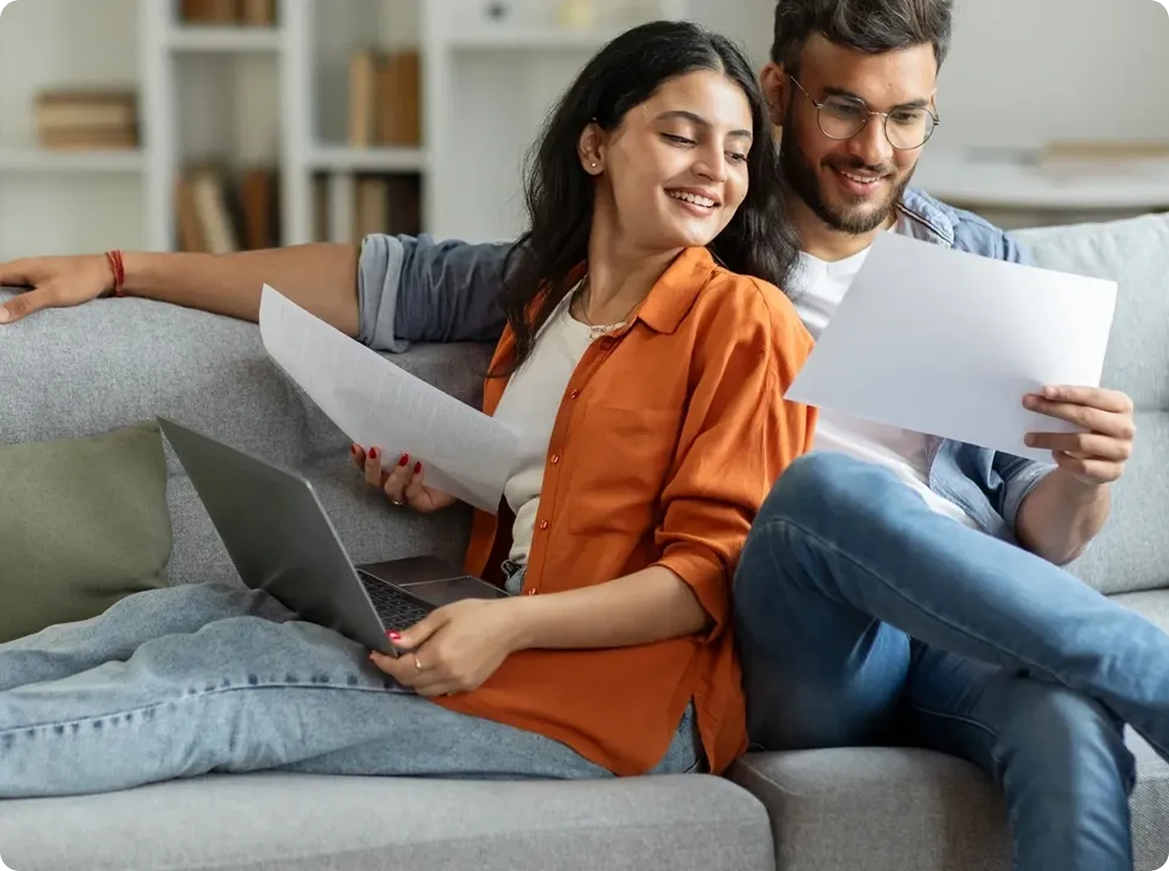 A man and woman sit on a couch together, looking at papers and a laptop, appearing to review documents at home.