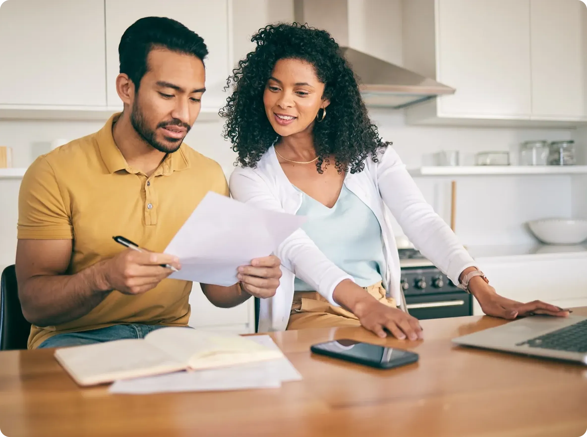 A man and woman sit in a kitchen, reviewing documents together with a notebook and smartphone on the table in front of them.